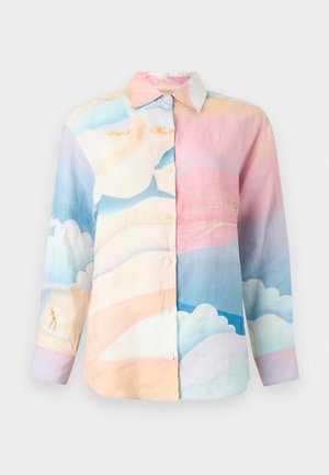 Personne portant des lunettes de soleil, une chemise à manches longues multicolore pastel, un jean bleu raccourci, des chaussettes blanches et des mocassins noirs, debout les mains dans le dos.