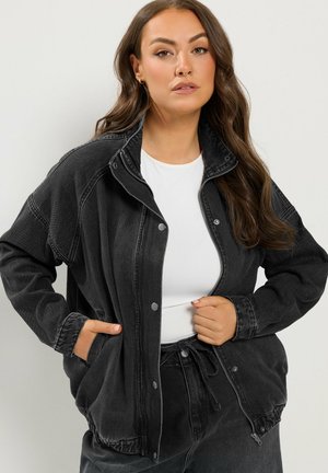 Femme aux longs cheveux bruns portant une veste en jean noire oversize par-dessus une chemise blanche, debout avec une main dans la poche et l'autre tenant la veste.