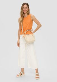 Beige handtas met een gestreepte handgreep, gedragen met een oranje mouwloos topje en witte wijde broek, met beige sandaaltjes met bandjes.