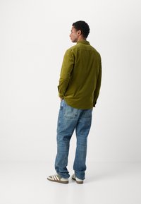 Cămașă din catifea cord cu guler, mâneci lungi și tiv rotunjit, de culoare verde măsliniu. Asortată cu pantaloni din denim de culoare albastru deschis și teniși bej cu dungi.