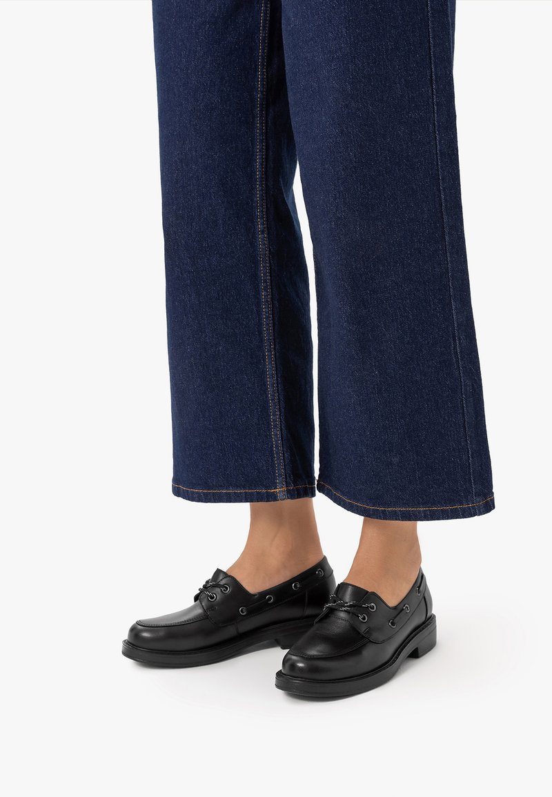 Personne portant des mocassins en cuir noir avec des détails en dentelle et un jean denim bleu foncé à jambes larges, debout sur une surface blanche.