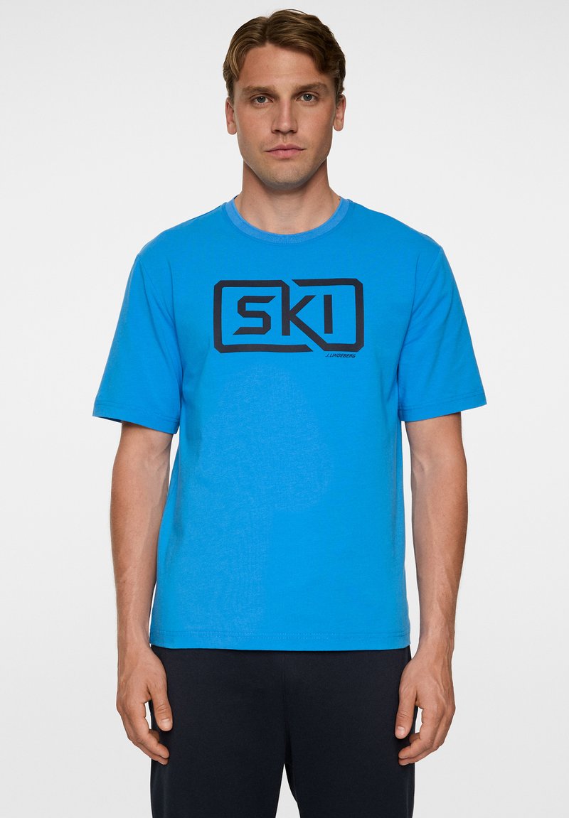 Sininen puuvillapaita, jonka pyöreä pääntie ja lyhyet hihat. Edessä on näyttävä musta "SKI" -logo. Sujuva rakenne.