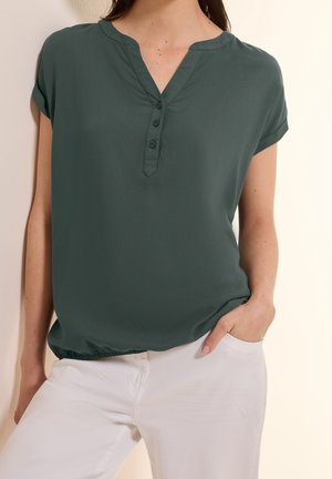 Vrouw draagt een donkergroene blouse met korte mouwen met knopen bij de hals en een witte broek, staand met één hand in de zak.