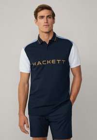 Navyblaues und weißes Polohemd mit kurzen Ärmeln und Kragen, mit goldfarbenem "HACKETT"-Logo über der Brust und kontrastierendem Schulterdesign.