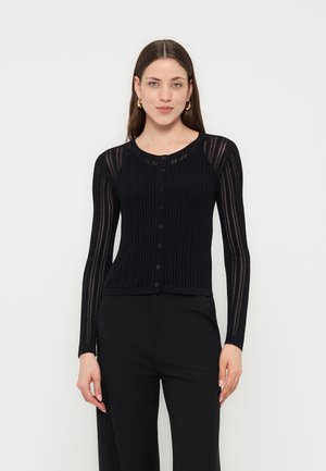 Frau trägt eine schwarze, langärmelige, geknöpfte Strickjacke mit vertikalen, offen gestrickten Streifen, kombiniert mit schwarzen Hosen vor weißem Hintergrund.