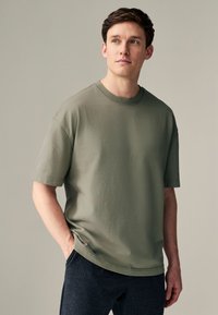 Olivengrøn bomulds-T-shirt med rund halsudskæring, korte ærmer og en løs pasform, kombineret med mørkegrå bukser. Glat tekstur, enkelt design.