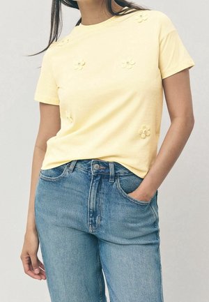 Frau trägt ein hellgelbes Kurzarmshirt mit kleinen Blumenapplikationen und blaue hoch taillierte Jeans, eine Hand in der Tasche.
