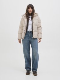 Beige pufjakke med hætte, der har trykknaplukninger og en quiltet tekstur, parret med lyseblå brede jeans og brune sko.