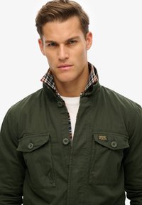 Chaqueta verde oscura con dos bolsillos en el pecho, cierre de botón y un forro de cuadros visible en el cuello. La tela parece ligera y texturizada.