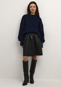 Pull en maille bleu marine avec manches surdimensionnées, mini-jupe en cuir noir et bottes noires montant jusqu'aux genoux. Texture lisse, coupe décontractée et taille froncée.
