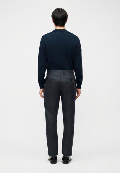 Man die een zwart shirt met lange mouwen, donkergrijze op maat gemaakte broek en zwarte nette schoenen draagt, staat met de rug naar de camera tegen een effen witte achtergrond.