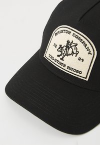 Crna kapa sa strukturiranim krojem, s kremastom oznakom. Oznaka prikazuje kauboja na kravi, uz natpise "Brixton Company" i "Tri-State Rodeo."