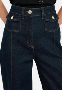 Tmavě modré džíny z denimu s vysokým pasem, dvěma předními kapsami a kontrastním žlutým prošíváním. Vybaveny kovovým knoflíkem a rovnou nohavicí.