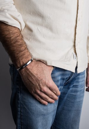 Srebrna kovinska zapestnica na zapestju, gladka površina, krožna oblika, nošena z svetlim srajco na gumb in modrimi jeans hlačami.