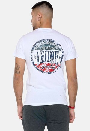 Mann mit kurzen Haaren und Tattoos, der ein weißes T-Shirt mit großem, abgenutztem Leone Italy-Logo auf dem Rücken und dunkelgraue Hose trägt.