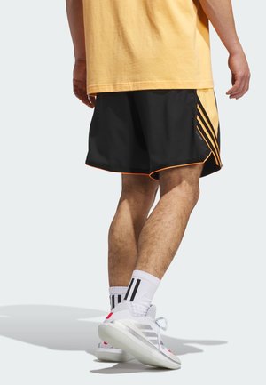 Un short de sport noir avec des accents orange et trois rayures verticales, associé à des baskets blanches et des chaussettes rayées, porté par une personne.