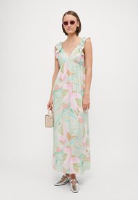 Μάξι φόρεμα σε ανοιχτό πράσινο χρώμα με λουλουδάτο μοτίβο σε ροζ και μπεζ. Διαθέτει ριγμένες τιράντες και εφαρμοστό μέσο. Το μοντέλο κρατάει μια μικρή τσάντα.