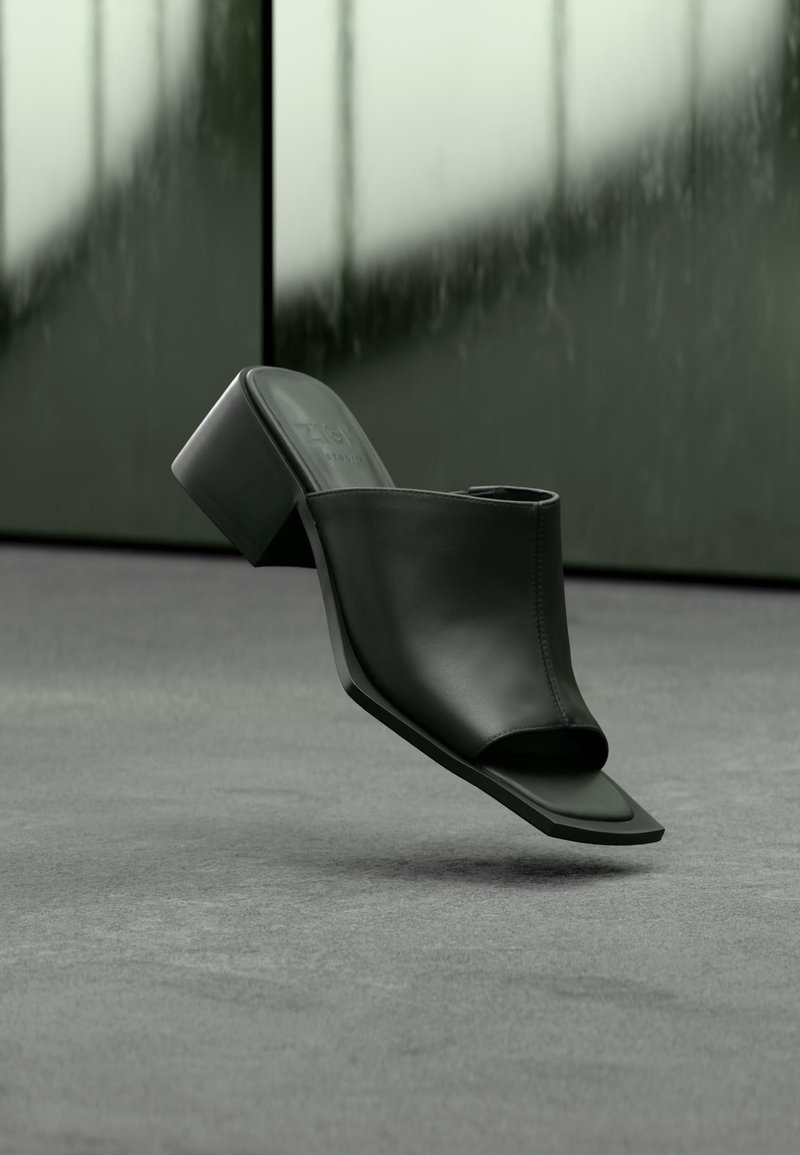 Μαύρο δερμάτινο mule με τετράγωνη ανοιχτή μύτη και χοντρό τακούνι να αιωρείται πάνω από γκρι χαλί σε μινιμαλιστικό εσωτερικό περιβάλλον.