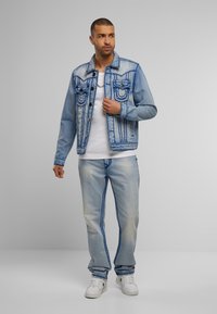 Denimjack met blauwe stiksels, twee voorkauzen; gedragen met lichtblauwe jeans. Witte t-shirt eronder en witte sneakers.
