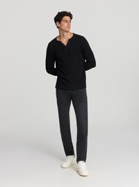 Maglietta nera a maniche lunghe con scollo alla henley parzialmente abbottonato, abbinata a jeans grigio scuro e sneakers bianche. Il modello è in piedi con le mani dietro la schiena.