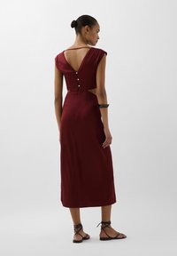 Burgundy midi kjole med korte ærmer, udsnit bagpå og knapdetaljer. Glat tekstur, tætsiddende silhuet, parret med sorte spændte sandaler.