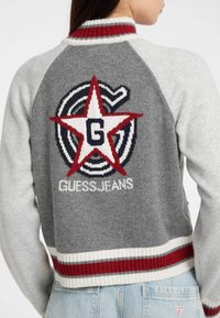 Giacca bomber grigia lavorata a maglia con strisce rosse e bianche. Presenta un grande logo a stella rossa con il testo "GUESSJEANS" sul retro. Design corto.