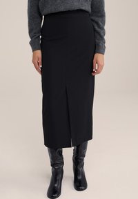 Fustă midi neagră dintr-un material elastic, cu o fanta în față, asortată cu cizme din piele neagră până la genunchi. Creată cu o siluetă ajustată.
