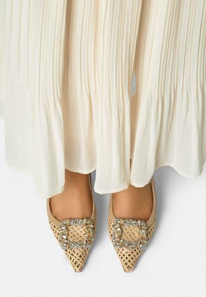 Beige gevlochten platte schoenen met puntige neus en grote rechthoekige en ronde strassversieringen, gedragen met een vloeiende crème plissé rok.