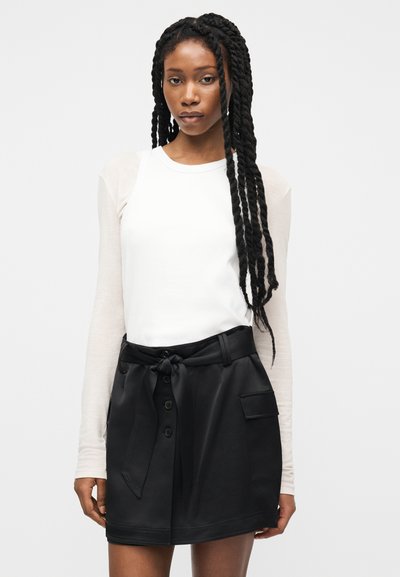 Νέα γυναίκα με μακριά πλεγμένα μαλλιά, ντυμένη με λευκό μακρυμάνικο τοπ και μαύρη φούστα με ζώνη, κουμπιά και τσέπη.
