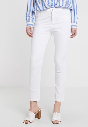 Woman wearing white slim-fit jeans, blue and white striped shirt, and white open-toe block heel sandals, standing against a plain background.