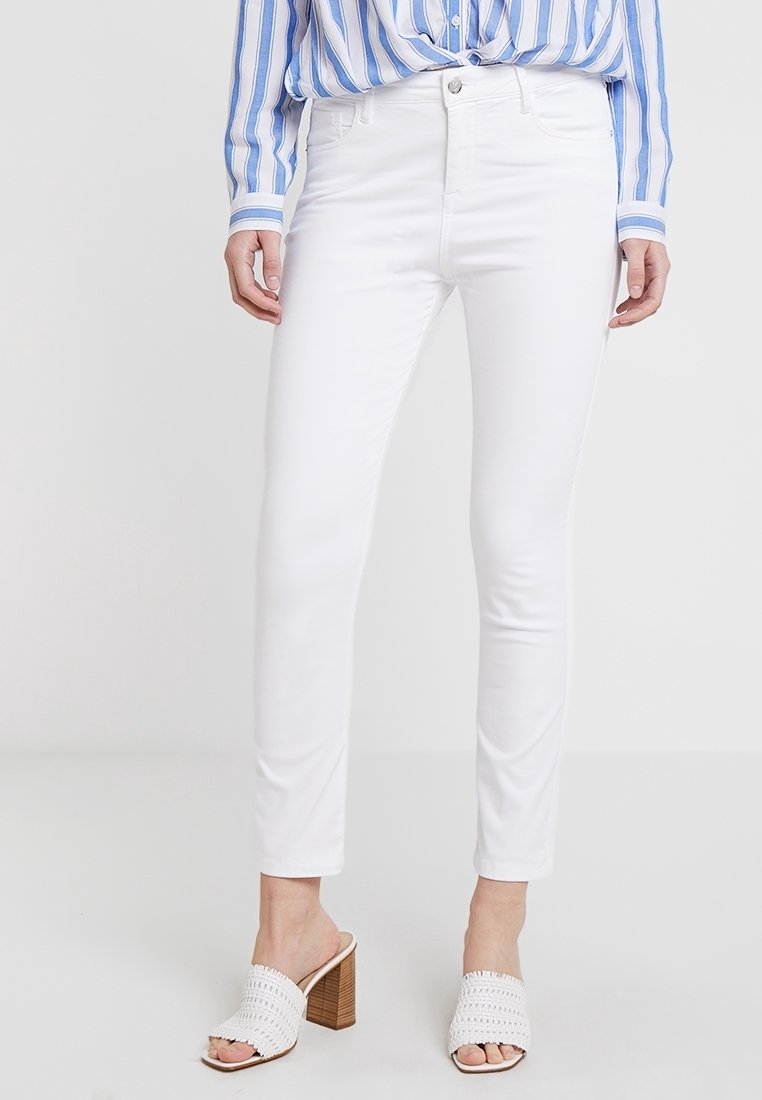 Femme portant un jean blanc ajusté, une chemise rayée bleu et blanc, et des sandales blanches à talons blocs ouverts, posant devant un fond uni.