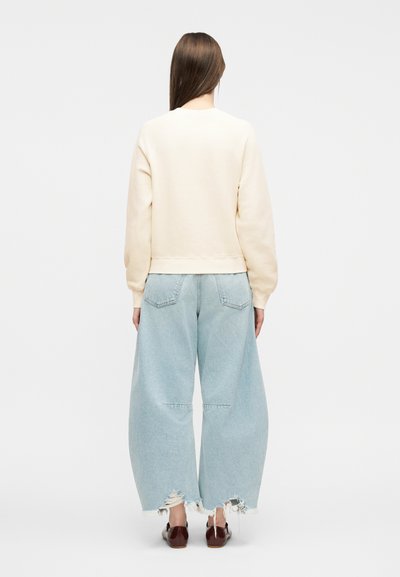 Γυναίκα που στέκεται πλάτη, φοράει κρεμ φούτερ, ανοιχτό μπλε τζιν παντελόνι με φαρδιά μπατζάκια και σκισίματα, και σκούρα καφέ παπούτσια χωρίς κορδόνια σε λευκό φόντο.