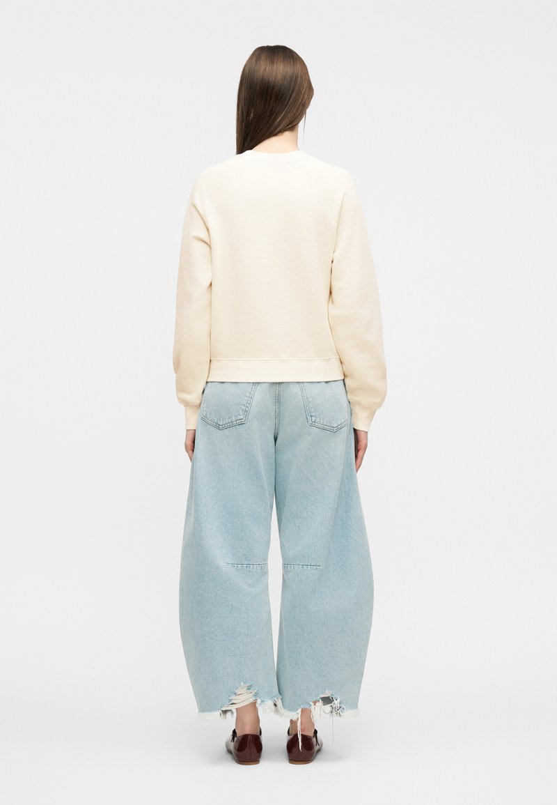 Femme debout de dos portant un sweat-shirt crème, un jean large bleu clair déchiré et des chaussures à enfiler marron foncé sur fond blanc.
