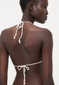 Bijela bikini gornja majica s podesivim vezicama na vratu i leđima, s ukrasima od perli za detalje. Glatka tekstura, minimalistički dizajn.