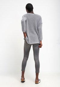 Vaaleanharmaa neulottu ylipitkä pusero, jossa on kolmen neljänneksen hihat, yhdistetty istuviin harmaisiin farkkuihin ja mustavalkoisiin sandaaleihin. Yksivärinen tausta.