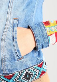 Käsi on pistetty sinisen farkkutakin sivutaskuun, jossa on värikkäästi kirjailtu hihansuu ja kuviollinen trimmi hemmen ympäri.