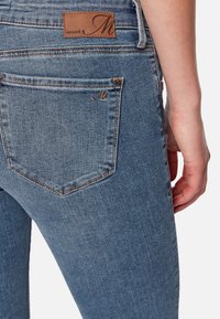 Vaqueros de mezclilla en azul claro con un parche de cuero marrón que presenta el logotipo de la marca. Textura desgastada con un diseño estándar de bolsillo trasero.