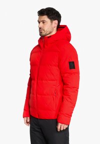 Red puffer jacket with a hood, featuring a zippered front, quilted texture, elastic cuffs, and a black logo patch on the left sleeve.