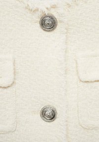 Witte textuurjack met twee voorkruiszakken en een grote decoratieve zilverkleurige knop met een cirkelvormig ontwerp. Zacht, geweven materiaal.