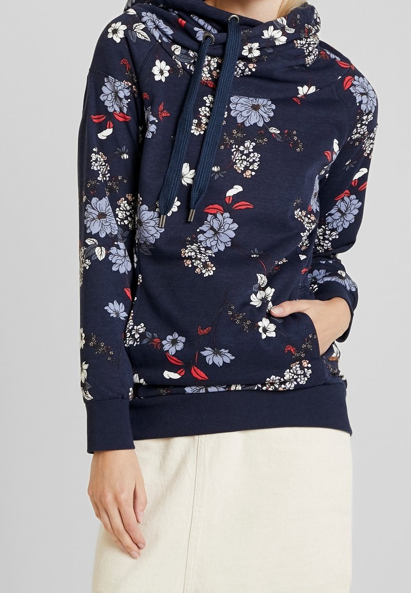 Sweat à capuche bleu marine à motifs floraux avec cordons, porté par une personne avec une main dans la poche avant et l'autre détendue le long du corps.
