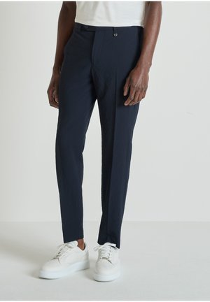 Uomo con pantaloni sartoriali blu navy e scarpe da ginnastica bianche, in piedi davanti a uno sfondo grigio chiaro uniforme.