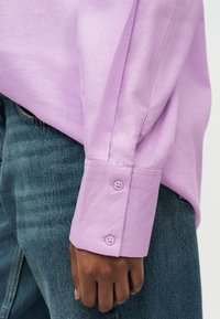 Mano de piel oscura descansando al lado, con puño de camisa lavanda abotonado y jeans de mezclilla azul contra un fondo liso.