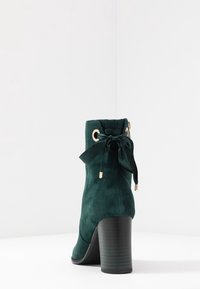 Stivale alla caviglia in suede verde con tacco a blocco, caratterizzato da occhielli dorati e un fiocco in tessuto sul retro, texture liscia e forma affusolata.