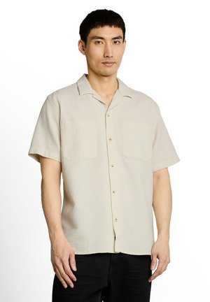 Man wearing a beige short-sleeve textured button-up shirt and black pants, standing against a white background.
