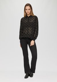 Zwarte doorschijnende shirt met grote zwarte stippen, knoopsluiting aan de voorkant en lange mouwen, gecombineerd met zwarte flared jeans en enkellaarsjes.