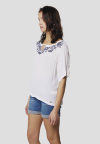 Blusa blanca con bordado floral en azul marino en el escote, mangas cortas y ajuste suelto, combinada con pantalones cortos de mezclilla. Tejido texturizado.