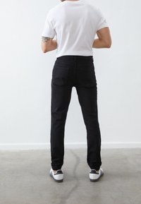 Jeans de mezclilla negros con corte ajustado, con bolsillos traseros y una textura suave, combinados con una camiseta blanca lisa y zapatillas negras y blancas.