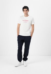 Weißes Baumwoll-T-Shirt mit rotem "Tommy Hilfiger"-Logo, kombiniert mit marineblauen Jogginghosen und weißen Sneakern. Schlichte, legere Sportbekleidung.
