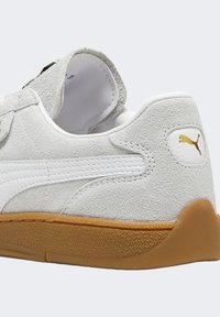 Białe zamszowe sneakersy z teksturowaną cholewką, gładkim białym paskiem ze skóry i gumową podeszwą. Zawierają małe złote logo na pięcie.