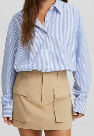 Femme portant une chemise boutonnée à rayures verticales bleues et blanches, rentrée dans une jupe beige avec de grandes poches avant.